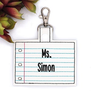 May include: A white rectangular keychain featuring a stitched design that mimics a sheet of lined notebook paper with a red vertical margin line and blue horizontal lines. The text Ms. Simon is embroidered in black on the front.