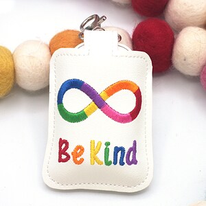 May include: A white leather keychain with a rainbow infinity symbol and the embroidered text "Be Kind." The keychain has a silver clasp and is surrounded by colorful felt balls.