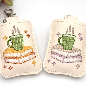 May include: Two cream-colored keychains with embroidered designs. Each features a green mug atop a stack of books, with steam rising and star accents. One has yellow books, the other purple.