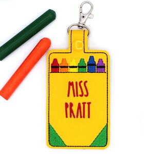 May include: Yellow crayon box keychain with a silver clasp, featuring a row of colorful crayons and the name "MISS PRATT" in red. Two chalk sticks, one green and one orange, are placed near the keychain.