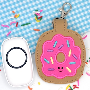 May include: A tan leatherette keychain with a pink donut design. The donut has a pink frosting with colorful sprinkles and a smiling face. The keychain has a silver-colored clasp.