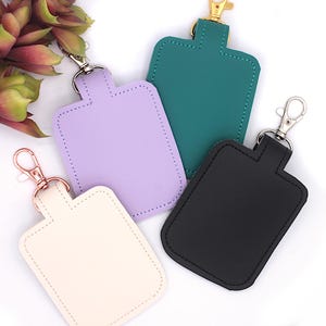 May include: Four rectangular keychains in cream, lavender, teal, and black. Each keychain has a stitched border and a metal clasp. The keychains are made of a smooth material and are designed to hold keys or other small items.