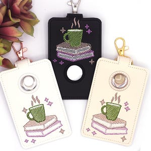 May include: Three keychains with a stitched design of a green mug on top of books. The keychains are black and off-white, with a silver or gold clasp. The design includes steam and star accents.
