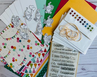 May include: A crafting kit with various elements for card making. Includes white envelopes, patterned paper with balloons and party hats, gnome stickers, colorful gems, twine, and a clear stamp set with birthday greetings.
