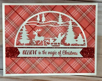 May include: A red and white plaid background with a white cutout of a winter scene. The scene includes a reindeer pulling a sleigh with Santa Claus, trees, and a snowy landscape. The text "BELIEVE in the magic of Christmas" is written on a red glitter banner.