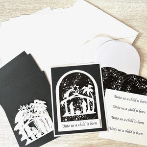 May include: A set of black and white stationery featuring a nativity scene with a star, palm trees, and figures. The cards include the text "Unto us a child is born" printed in black on white paper, accompanied by matching envelopes and circular inserts.