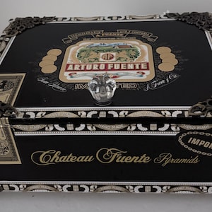 Caixa de charutos ARTURO FUENTE feita de madeira reaproveitada, com pés e cantos superiores em latão antigo. Puxador de vidro canelado. Forrada com feltro.