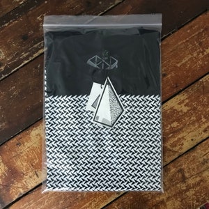 May include: A black and white t-shirt with a geometric pattern. The shirt is packaged in a clear plastic bag with a white tag that says "SHARE".