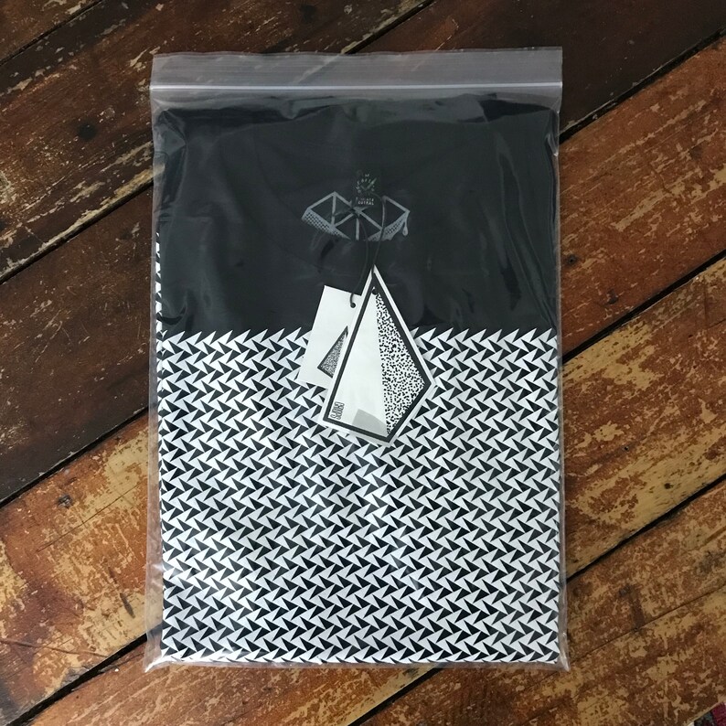 May include: A black and white t-shirt with a geometric pattern. The shirt is packaged in a clear plastic bag with a white tag that says "SHARE".