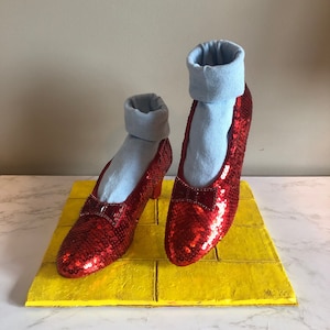 May include: A pair of red sequined shoes with blue fabric cuffs stand on a yellow brick road. The shoes have a pointed toe and a small bow on the front.