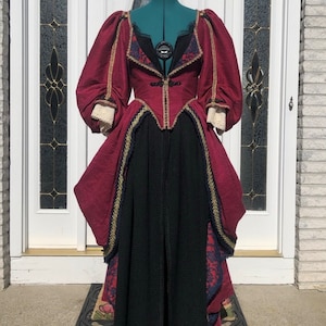 Puede incluir: Un vestido burdeos y negro con un corpiño ajustado, mangas abullonadas y una falda completa. El vestido tiene ribetes dorados y detalles de encaje.
