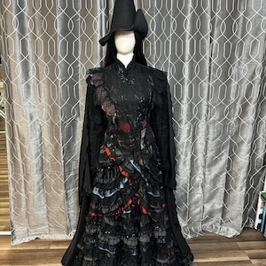 May include: A black witch costume with a tall, pointed hat and a long, flowing dress. The dress features layers of ruffles with red and blue accents. The costume is displayed on a mannequin.