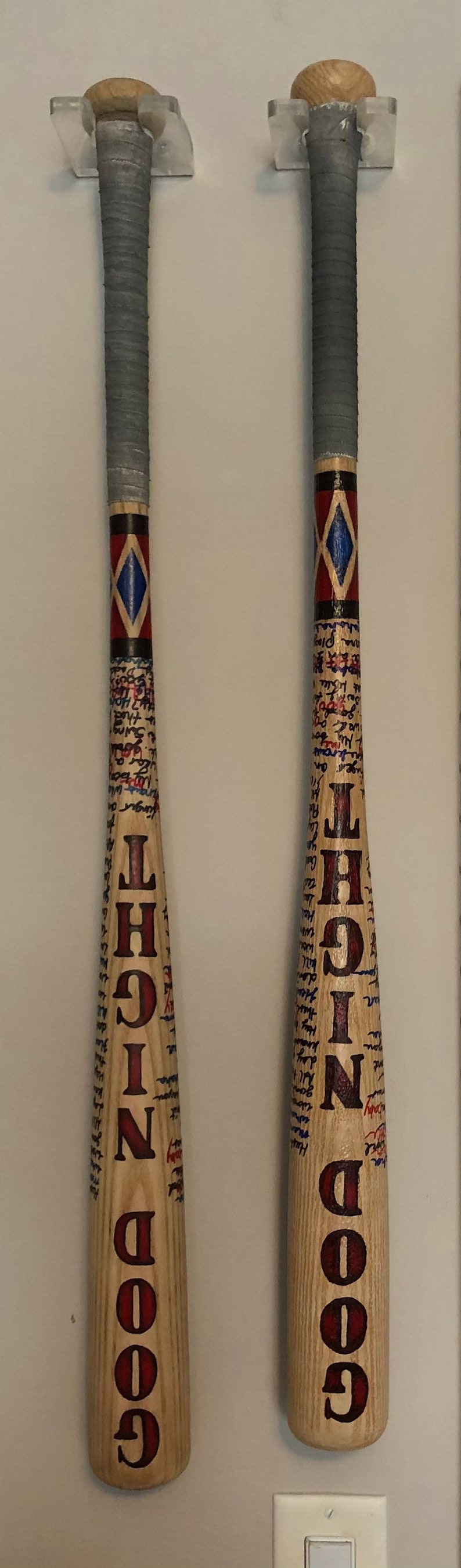 May include: Two wooden baseball bats with grey tape on the handle. The bats are painted with red and blue diamond patterns and have the words "GOOD NIGHT" written on them in red.