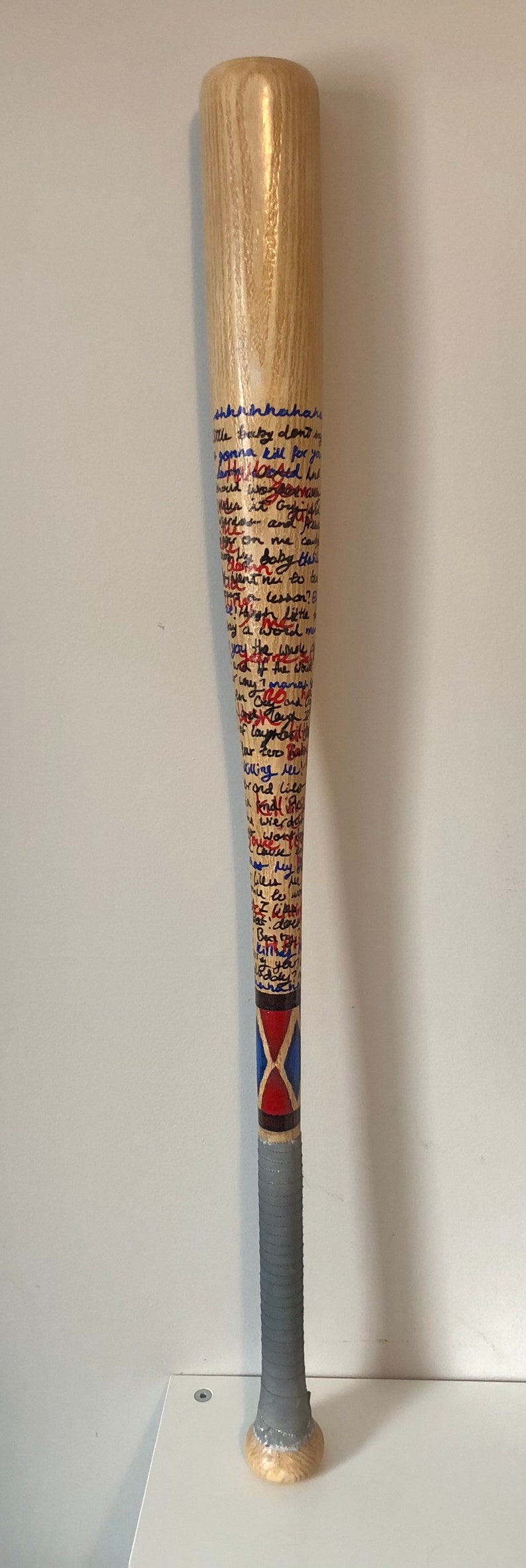 May include: A wooden baseball bat with a grey grip and red, blue and black painted accents. The bat is covered in handwritten text in blue and red ink. The text reads: "shhhinhahan ille baby don't gonna kill for yo phoned in wa houd os and on me ca Baby this went nu to tu Killing ree! Hond Lilo wierde My dou you"
