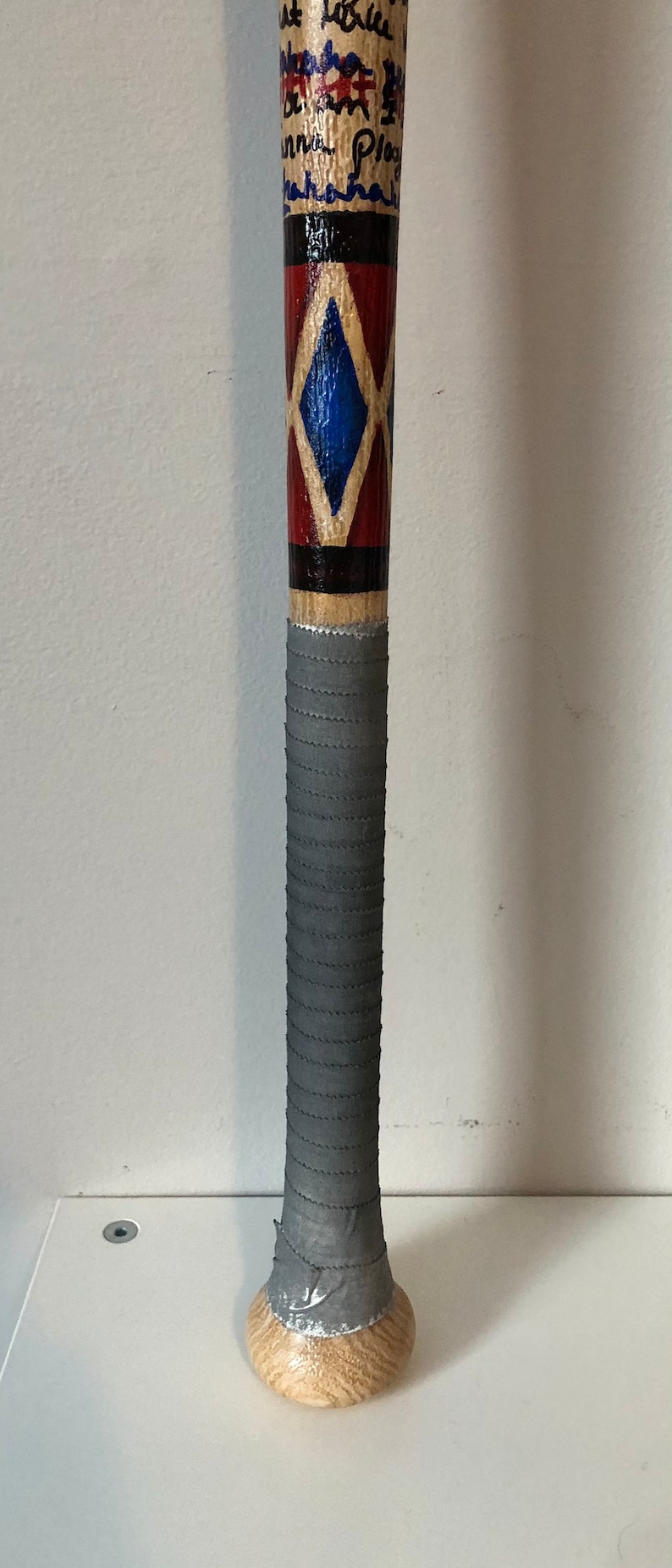 May include: A wooden baseball bat with a grey grip and a red, white and blue diamond pattern. The bat is painted with black and white writing.