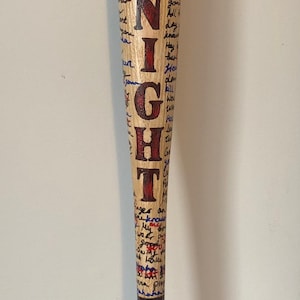 May include: A wooden baseball bat with the word "GOODNIGHT" written in red and black paint. The bat is covered in blue and red writing and has a blue and red diamond pattern near the bottom.