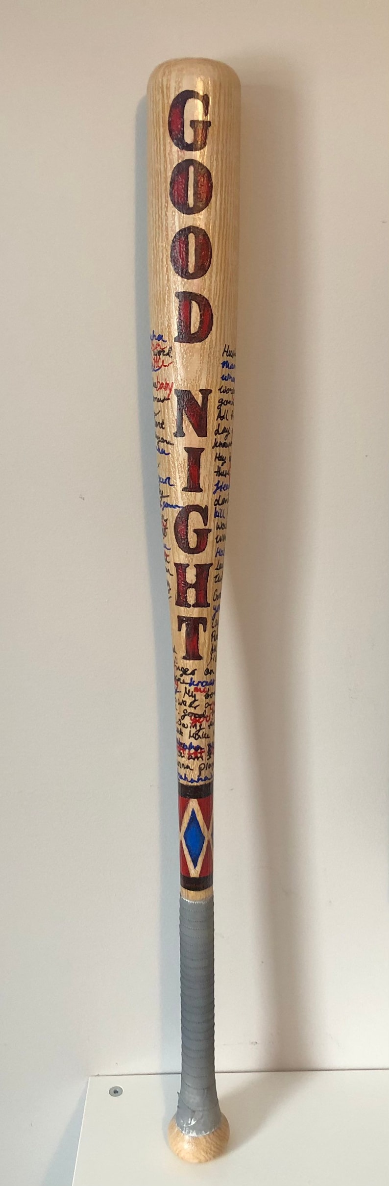 May include: A wooden baseball bat with the word "GOODNIGHT" written in red and black paint. The bat is covered in blue and red writing and has a blue and red diamond pattern near the bottom.