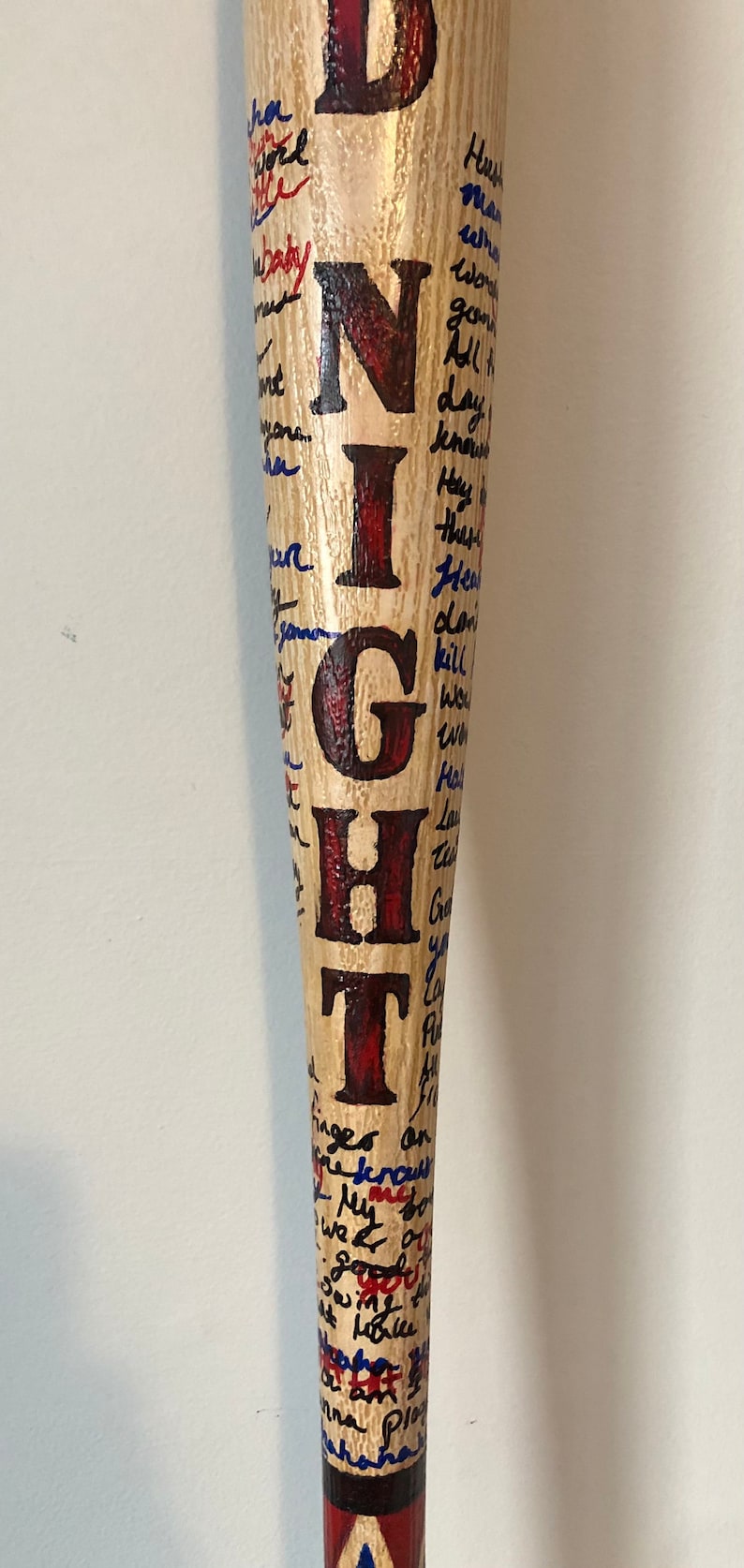 May include: A wooden baseball bat painted with the word "NIGHT" in red and blue letters. The bat is covered in handwritten messages in blue ink.