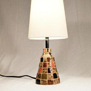 May include: A decorative table lamp with a white lampshade and a mosaic-tiled base. The base features a geometric pattern of brown, red, green, and black squares. The lamp has a black metal stem and a power cord.