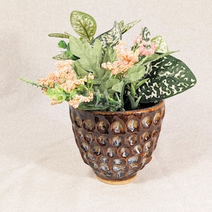May include: A brown ceramic pot with a textured, honeycomb-like pattern, filled with artificial plants. The arrangement includes green leaves, speckled leaves, and small pink flowers, creating a decorative display. The pot has a small, unglazed base.