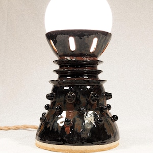 May include: A handcrafted ceramic table lamp with a dark brown, glossy finish. The lamp features a spherical white light bulb and a textured base with small, rounded embellishments. The lamp's cord is a natural, woven material.