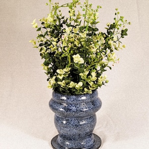 May include: A blue ceramic vase with a speckled pattern, holding a collection of green and white artificial plants. The vase has a bulbous shape with a round base and a decorative rim. The plants have small, delicate leaves and a few tiny pink buds.