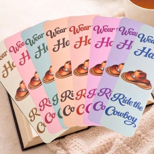 May include: A selection of colourful bookmarks featuring a cowboy hat design. The text "Wear Ho" or "Ride the Cowboy" is printed on each bookmark in a script font. The bookmarks are displayed over an open book.