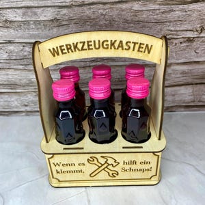 May include: A wooden carrier holding six small bottles with pink caps. The carrier has the words "WERKZEUGKASTEN" and "Wenn es klemmt, hilft ein Schnaps!" etched into the wood, along with a hammer and wrench graphic.