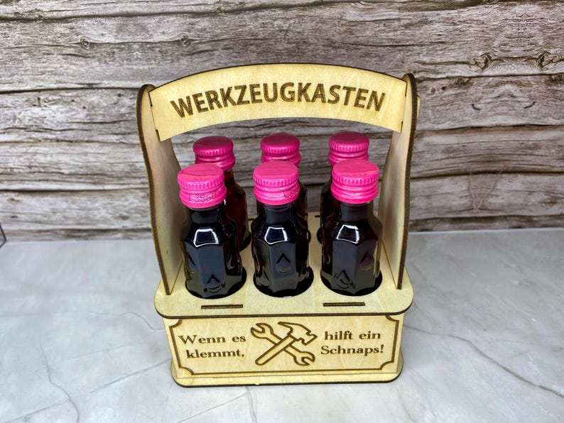 May include: A wooden carrier holding six small bottles with pink caps. The carrier has the words "WERKZEUGKASTEN" and "Wenn es klemmt, hilft ein Schnaps!" etched into the wood, along with a hammer and wrench graphic.
