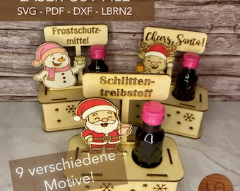 May include: Three wooden laser-cut display stands for miniature bottles, each featuring a festive character cutout of a snowman, a reindeer, or Santa Claus, with engraved German text labels and snowflake patterns on the base.