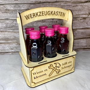 May include: A wooden carrier with six small bottles of dark liquid, each with a bright pink cap. The carrier has the words "WERKZEUGKASTEN" and "Wenn es klemmt, hilft ein Schnaps!" etched into the wood, along with a hammer and wrench design.