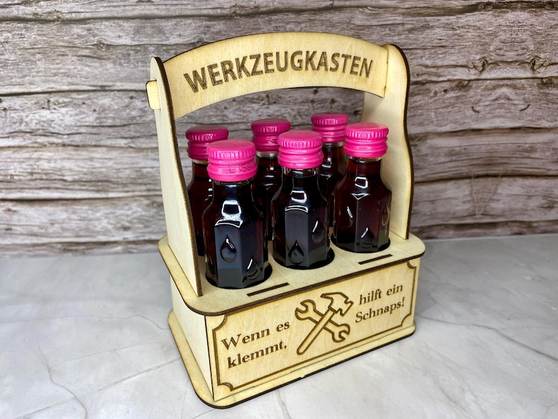 May include: A wooden carrier with six small bottles of dark liquid, each with a bright pink cap. The carrier has the words "WERKZEUGKASTEN" and "Wenn es klemmt, hilft ein Schnaps!" etched into the wood, along with a hammer and wrench design.