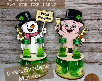 May include: Two laser-cut wooden bottle holders featuring a snowman and a pig, both wearing black top hats with green shamrocks. The snowman holds a sign reading Prost Neujahr. Each holder has two circular cutouts for small green glass bottles.