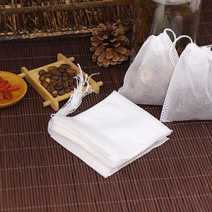 May include: A stack of white, square, muslin tea bags with a drawstring. The tea bags are on a brown bamboo mat.