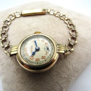 Op de afbeelding: Een gouden armbandhorloge met een ronde wijzerplaat en een gouden kettingarmband. De wijzerplaat heeft een witte achtergrond met zwarte Romeinse cijfers voor de uren en een blauwe secondewijzer. Op de wijzerplaat staat ook het woord "CENTRAL" in zwart gedrukt.