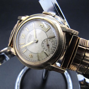 Peut inclure: Une montre-bracelet vintage dorée avec un cadran crème et des aiguilles dorées. La montre porte l'inscription "Revere De Luxe" et un petit cadran secondaire. Elle possède un bracelet doré et est présentée sur un support argenté.
