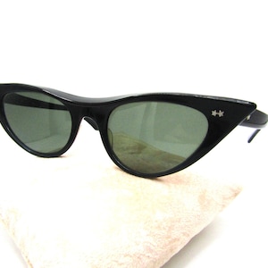 Puede incluir: Gafas de sol negras de ojo de gato con lentes verdes y detalles plateados. Las gafas de sol están apoyadas sobre una superficie de tela marrón claro.