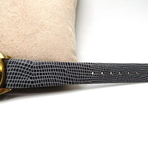 K&ouml;nnte beinhalten: Eine goldfarbene Armbanduhr mit einem schwarzen Lederarmband. Das Zifferblatt der Uhr zeigt Tag und Datum an, und das Armband hat ein strukturiertes Muster.
