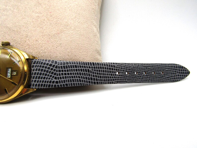 K&ouml;nnte beinhalten: Eine goldfarbene Armbanduhr mit einem schwarzen Lederarmband. Das Zifferblatt der Uhr zeigt Tag und Datum an, und das Armband hat ein strukturiertes Muster.