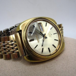 May include: A gold-toned Seiko automatic wristwatch with a square face and a metal band. The watch face has black hour and minute hands, a black second hand, and a date window that displays "FRI 21".