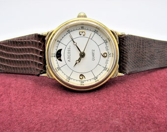 May include: A gold-tone watch with a white face and a brown leather band. The watch has a moon phase complication and the text "AXCESS" and "QUARTZ" on the face.
