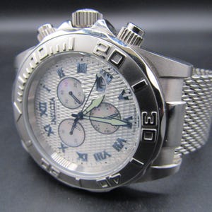 May include: A silver-tone stainless steel chronograph watch with a textured white dial, Roman numeral hour markers, and a mesh metal strap. The watch features three sub-dials, a date window, a rotating bezel with engraved numbers, and the brand name Invicta.