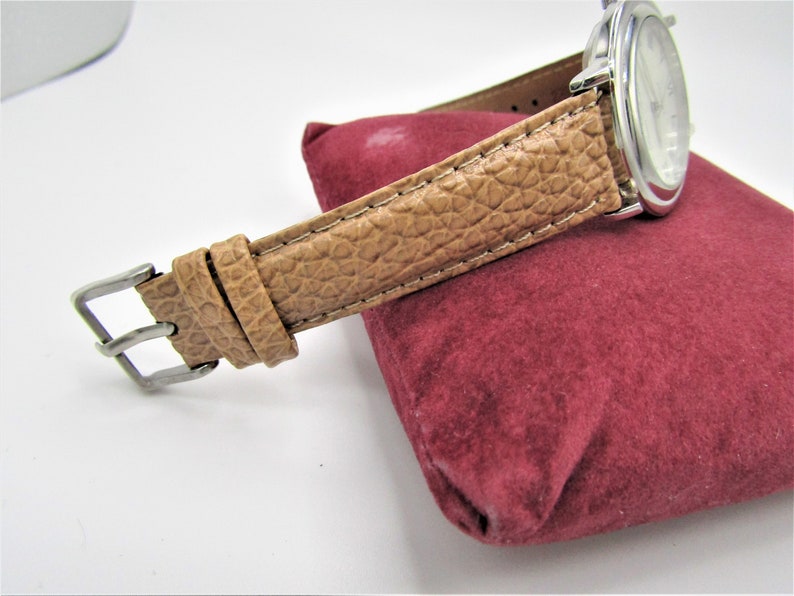 May include: A silver watch with a brown leather band. The watch is on a red velvet cushion.