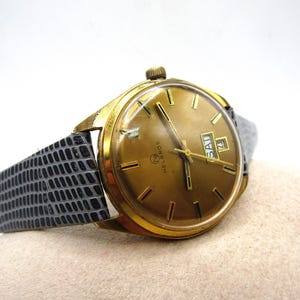 K&ouml;nnte beinhalten: Eine goldfarbene Armbanduhr mit einem schwarzen Lederarmband. Das Zifferblatt der Uhr ist goldfarben mit schwarzen Markierungen und einem Wochentags- und Datumsfenster. Die Marke der Uhr ist "Melbros".
