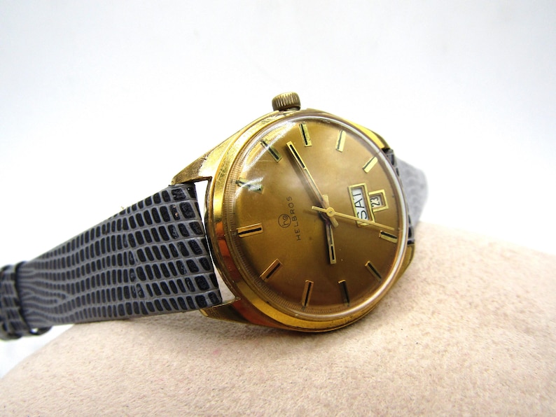 K&ouml;nnte beinhalten: Eine goldfarbene Armbanduhr mit einem schwarzen Lederarmband. Das Zifferblatt der Uhr ist goldfarben mit schwarzen Markierungen und einem Wochentags- und Datumsfenster. Die Marke der Uhr ist "Melbros".