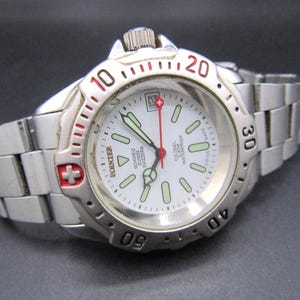 May include: A silver-tone stainless steel wristwatch with a white dial, luminous hour markers, and a red second hand. The bezel features black numerals and a red Swiss cross emblem. The watch includes a date window and a metal link bracelet.