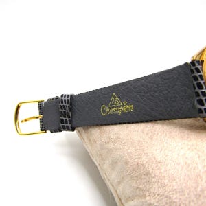 K&ouml;nnte beinhalten: Eine goldfarbene Armbanduhr mit einem schwarzen Lederarmband. Das Armband tr&auml;gt die Worte "Champion" in Pr&auml;gung.