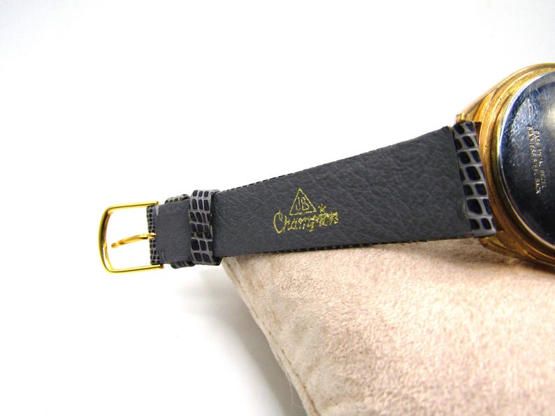 K&ouml;nnte beinhalten: Eine goldfarbene Armbanduhr mit einem schwarzen Lederarmband. Das Armband tr&auml;gt die Worte "Champion" in Pr&auml;gung.