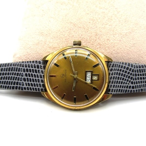 K&ouml;nnte beinhalten: Eine goldfarbene Armbanduhr mit einem grauen Lederarmband. Das Zifferblatt der Uhr hat einen goldenen Hintergrund mit schwarzen Markierungen und ein Datumsfenster, das "SAM 23" anzeigt. Die Marke der Uhr ist "Helbros".