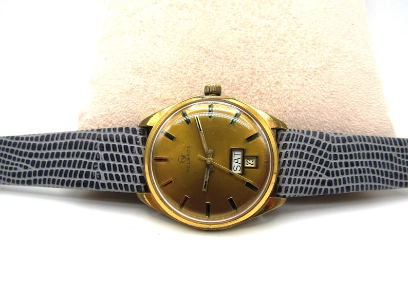 K&ouml;nnte beinhalten: Eine goldfarbene Armbanduhr mit einem grauen Lederarmband. Das Zifferblatt der Uhr hat einen goldenen Hintergrund mit schwarzen Markierungen und ein Datumsfenster, das "SAM 23" anzeigt. Die Marke der Uhr ist "Helbros".